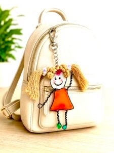 Bag Charm from the Mary-Anne Collection showing Cheeky Girl Character in an orange dress made with uv resin  and hair tied in bunches attached to a small ladies ruck sack in cream colour 