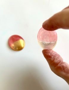 two resin pieces that have been coloured using mica powder one held up showing how translucent the colours look when held to the light compared to the one that is flat on surface showing bright bold red and gold colouring